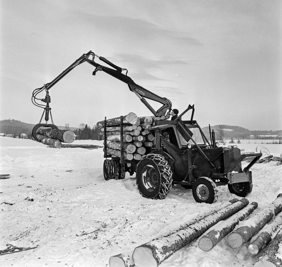 Tømmerkjøring med traktor, muligens på Jømna i Elverum kommune i Hedmark vinteren 1969.  Traktorer med tømmertilhenger og griplasterkran ble lansert som en nyhet i slutten av 1960-åra.  Da hadde den svenske produsenten HIAB lansert sin «skogselefant».  De norske skogteknologene var skeptiske til denne maskintypen.  Med den norske eiendomsstrukturen og i norsk terreng var det de rammestyrte helstammelunnerne som var framtidsrettet, hevdet de.  Mjøsen, Glommen og Nedre Glommen skogeierforeninger satte likevel i gang forsøk med det de kalte «kombinasjonsmaskiner» - traktorer med griplastere og boggidoninger.  I slutten av 1960-åra kunne norske maskinleverandører tilby flere varianter av slike maskiner.  Den svenaske «skogselefanten» med HIAB-kran på en rammestyrt traktor var den mest avanserte, men det ble også levert svenske og finske krantyper som kunne monteres på kraftige landbrukstraktorer.  Også den norske maskinfabrikken Hymas i Brumunddal bygde tidlig ei gripeklo som kunne monteres i steden for skuffa på sine traktorgravere, som allerede var mye brukt i det norske skogbruket.  Dermed kunne man få helårsdrift på en traktor som inntil da i hovedsak hadde vært brukt sommerstid.  Som tømmerlastingsmaskin var Nuffieldene med kraner fra Hymas mest anvendelige på velteplasser, men de kunne også trekke tilhengere med tømmer som var pålesset ved hjelp av traktorkrana.  Det er usikkert om det vi ser på fotografiet er et Hymas-produkt, eller om det er en svensk modell.  

Etter hvert måtte de rammestyrte stammelunnerne vike plassen for «kombinasjonsmaskiner» med kran og tilhenger for tømmerlass.  Skogteknologene gav dem etter hvert betegnelsen «lastetraktorer», mens maskinforhandlerne brukte betegnelsen «lassbærere», for å tydeliggjøre at det tømmeret man fikk fram ikke ville være sølete og tilgriset, slik stammelunnet virke kunne være, i hvert fall i sommerhalvåret.