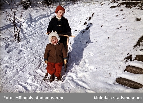 Två barn med en spark i snön. Det ena barnet kör och det andra åker med. 
Okända barn, plats och årtal.