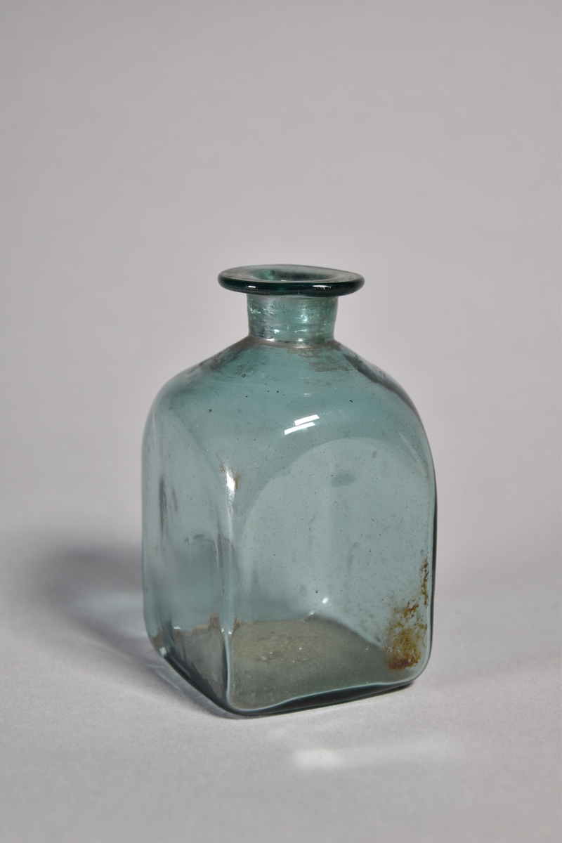 Flaska av gröntonat glas. Kvadratisk med kort hals och utvikt mynning.
