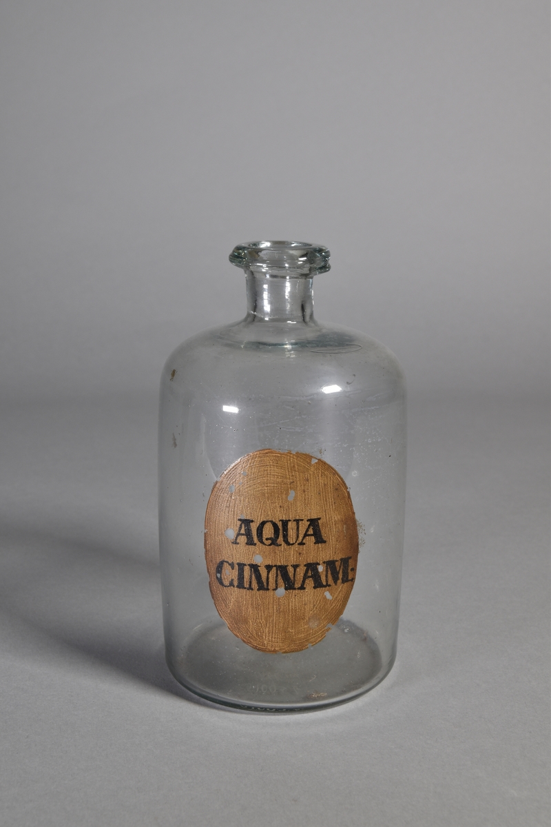 Flaska av klart glas, cylindrisk, kort hals med utsvängd mynning. Påmålad etikett med svart text. Glaset iriserat.