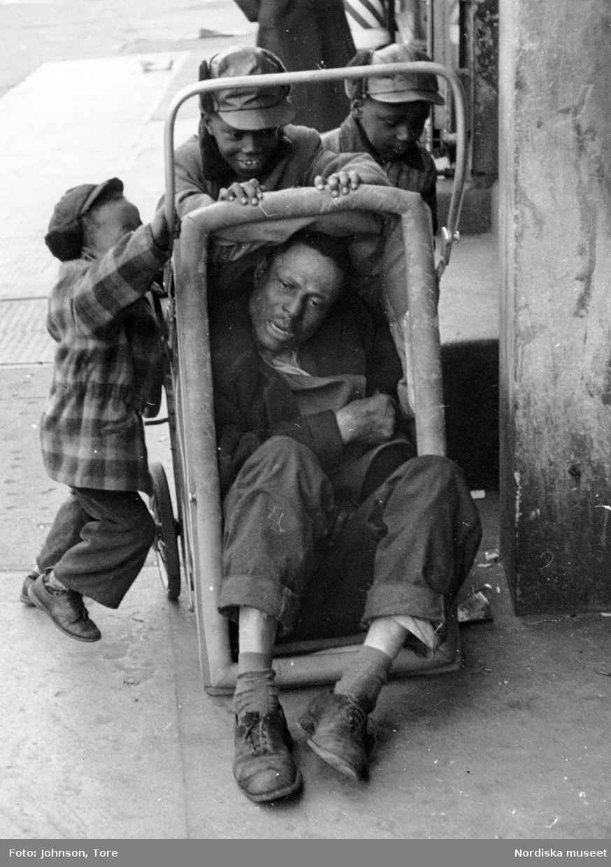 En utslagen man ligger i en barnvagn som knuffas av en grupp barn. Lenox Avenue, Harlem, New York, USA.