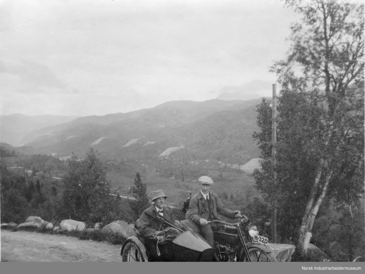 Motorsykkelist med passasjer i sidevogn. Steintippene nedover langs anlegget på Rjukan vises tydelig i fjellsiden.