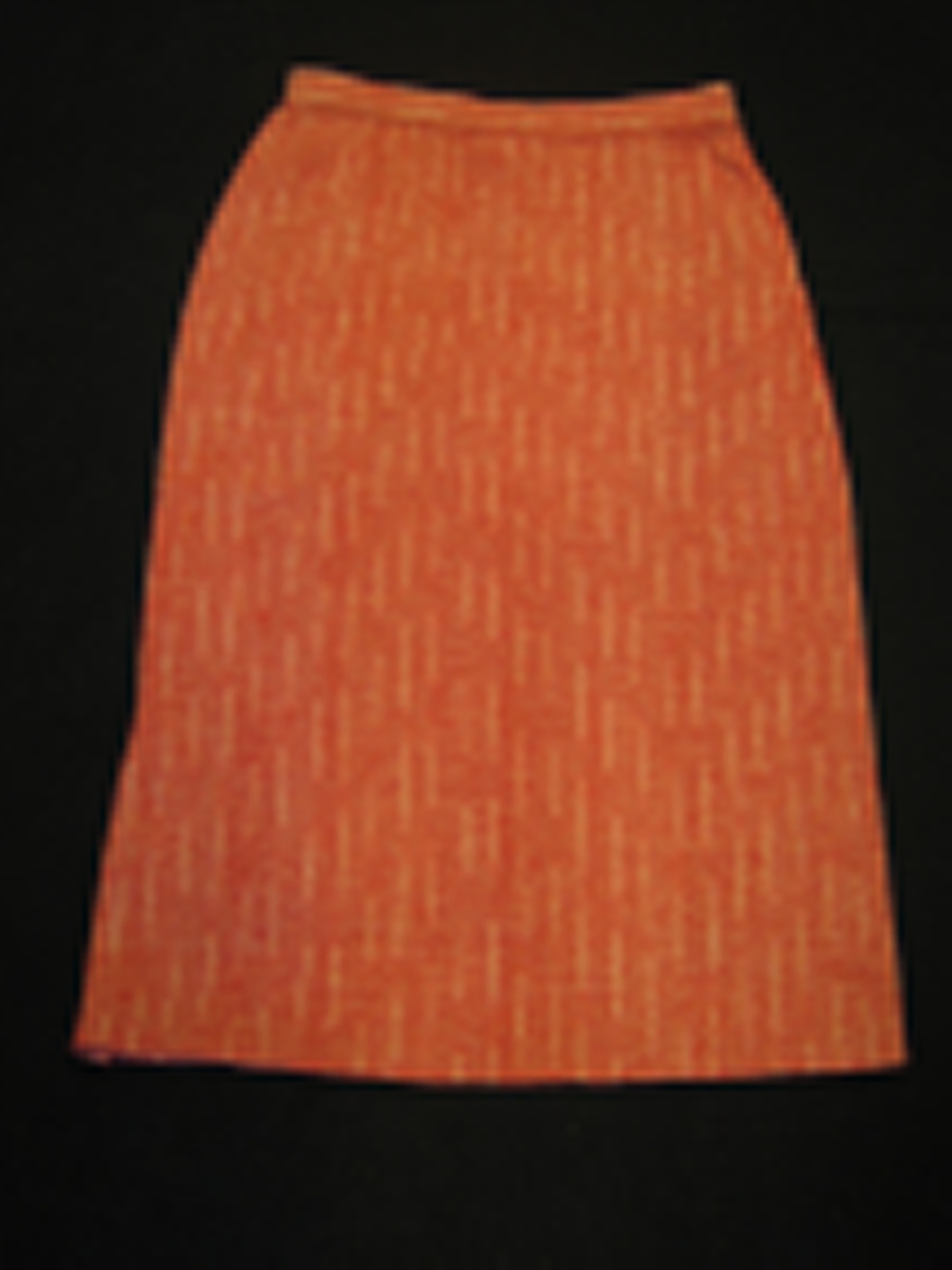 Rädvit randig rak kjol i ull. Tyget är vävt i kypert. Kjolen har två sprängveck bak och en 25 mm bred linning med knapp, knapphå och dragkedja i vänster sida. Kjolen är försedd med ett 19 cm långt gångveck i vänster sida.