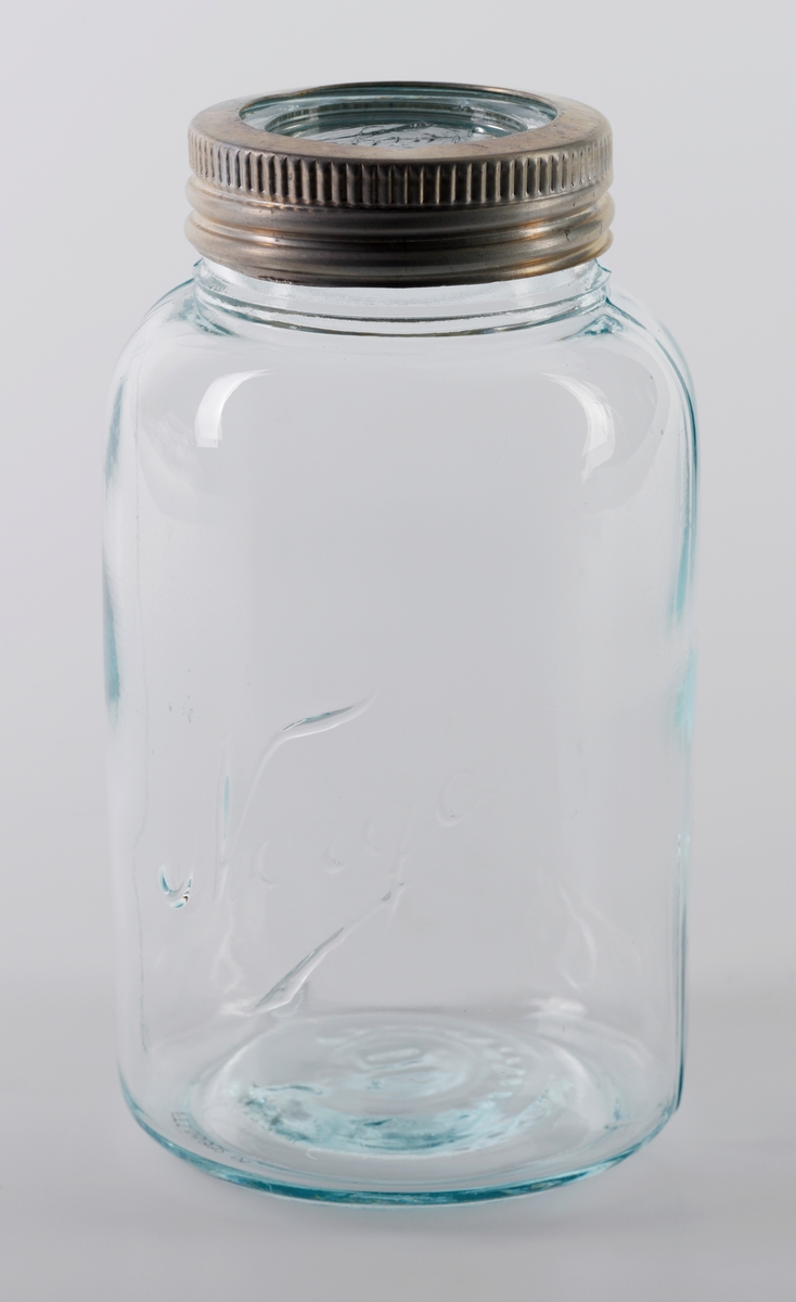 Tre "Norgesglass" med løst glasslokk og mansjett laget av aluminium til å skru fast lokket. Glasset er blankt med grønnskjær i. På lokket og på siden av glasset er det skrevet "Norge" i relieff.