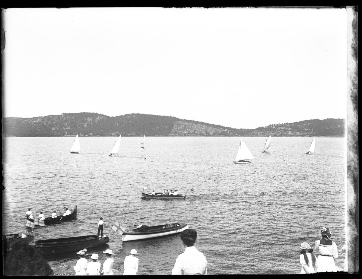 Kappseglingstävling på Mjörn fotograferat från startplatsen vid Lövekulle. Åskådare sitter i båtar och står på stranden medan ett antal segelbåtar tävlar ute på sjön.