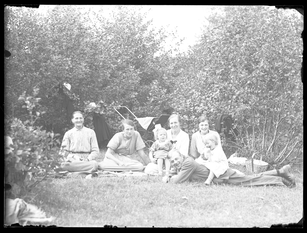 John Hjelm med sin familj har picknick i gräset mellan träden. Sju personer syns på bilden, bakom dem står en barnvagn och i trädgrenarna har de hängt av sig jackor och hattar.