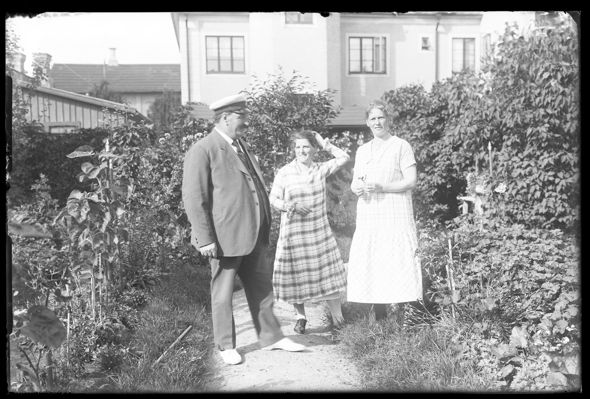 En man i kostym, studentmössa och ljusa tygskor samt två kvinnor i rutiga sommarklänningar står på en grusgång i en lummig trädgård. I bakgrunden syns ett hus. I fotografens anteckningar står det "Fam. Wincrantz". Bilden är tagen i samband med herr och fru Wincrantz 50årsdag.