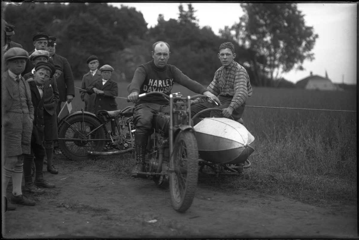 Harald Karlborg fotograferad i samband med Alingsås Motorklubbs backtävlan. Karlborg sitter på en motorcykel med sidovagn, i vagnen sitter en pojke. Bredvid dem står åskådare.
