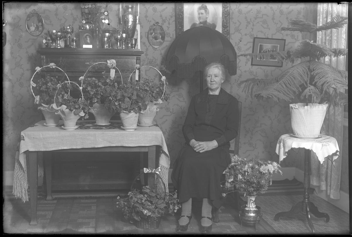 Fru Hanson sitter vid ett bord, omgiven av blommor, i samband med sin 60årsdag. I bakgrunden står en byrå med pokaler ovanpå.