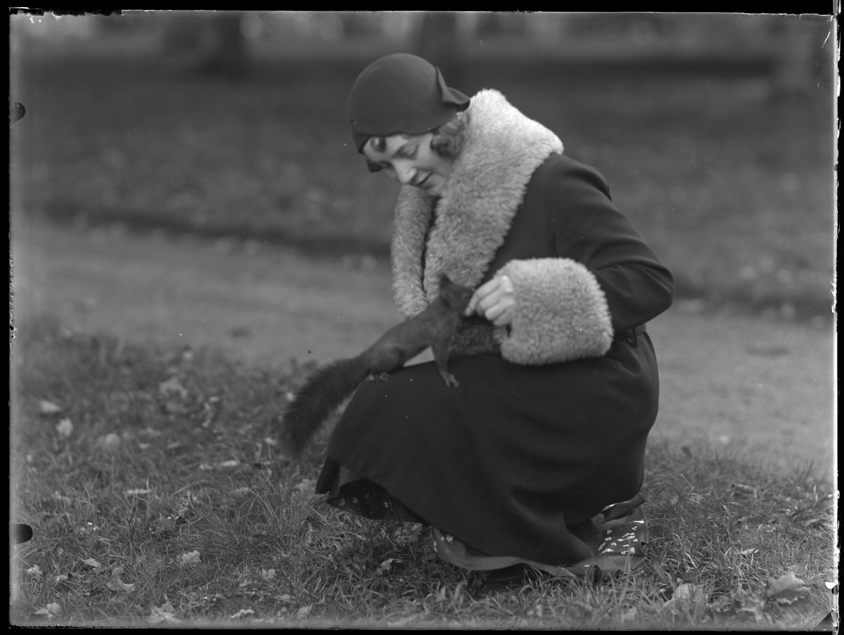 Gulli Svensson, i pälskantad kappa, fotograferad i en park när hon sitter på huk med en ekorre i knät.