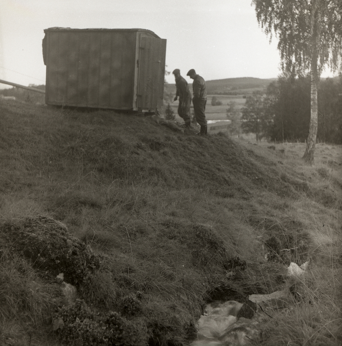 Två män står i en backe nedanför en koja, 25 augusti 1956.
