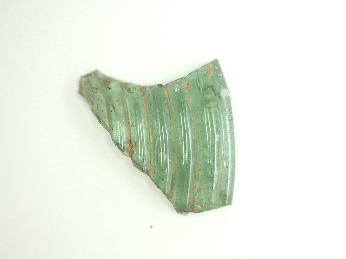 Fragment av foten till en remmare eller ett så kallat nuppenglas i grön glasmassa (se referenser/digitala objekt). Foten är troligen pressad med koncentriska räfflor på över- och undersidan. Passar ihop med M 17000-629 b.