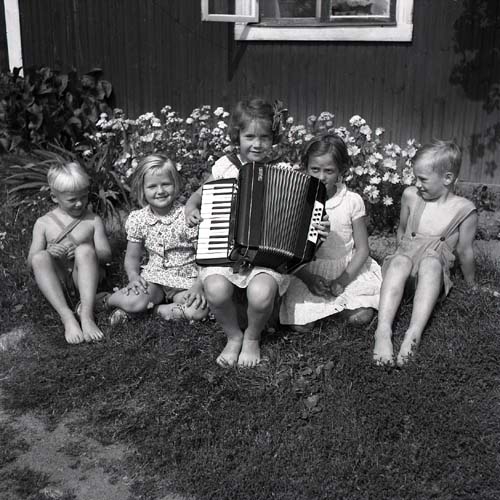 Fem barn sitter på en gräsmatta och en flicka spelar på ett dragspel, 1947.