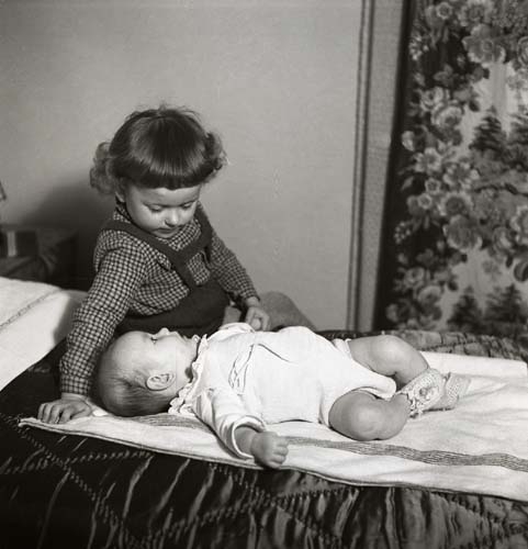 Två barn befinner sig på en säng. Det yngre barnet ligger på rygg iförd sparkdräkt medan det äldre barnet sitter intill och håller det yngre i handet, 1953.