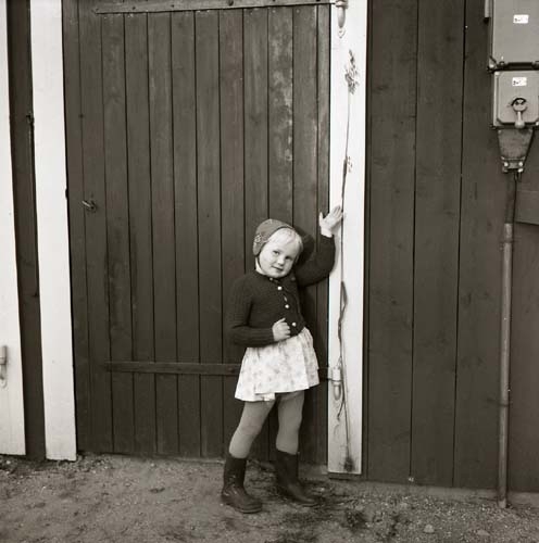 En flicka iförd kofta, mössa, kjol och stövlar poserar intill en dörr, 1956.