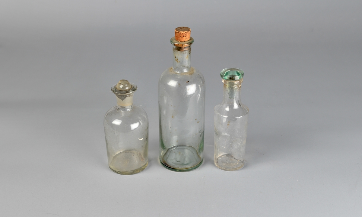 Rette og runde flasker i glass med innsnevring til halsen. Rundt hals. A og C har glasskork, B har kork. A har korken delvis knust på toppen.