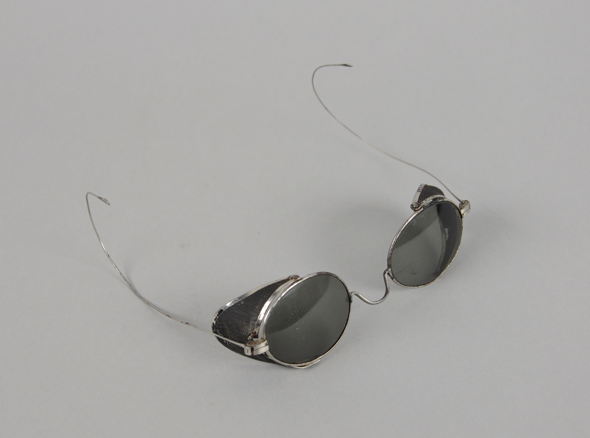 Ovale solbriller med metallinnfatning og beskyttelse på yttersidene av brilleglassene. Smale stenger med store bøyler ytterst.