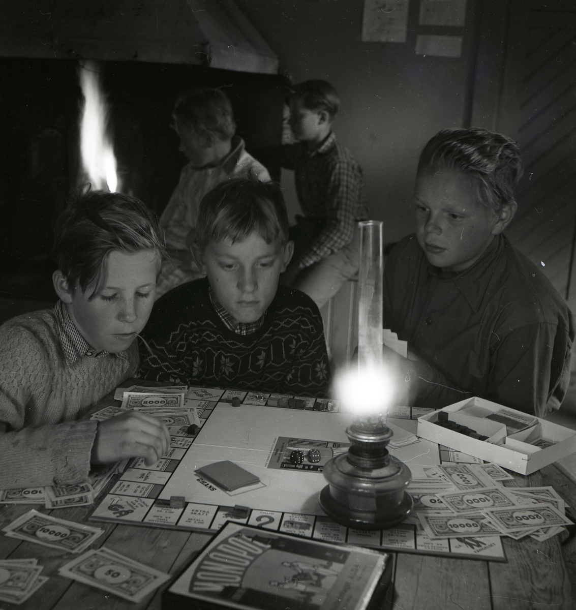 Tre pojkar sitter vid ett bord och spelar Monopol i skenet av en fotogenlampa. I bakgrunden syns en öppen spis vid vilken två pojkar står, Norrvåga 1953.