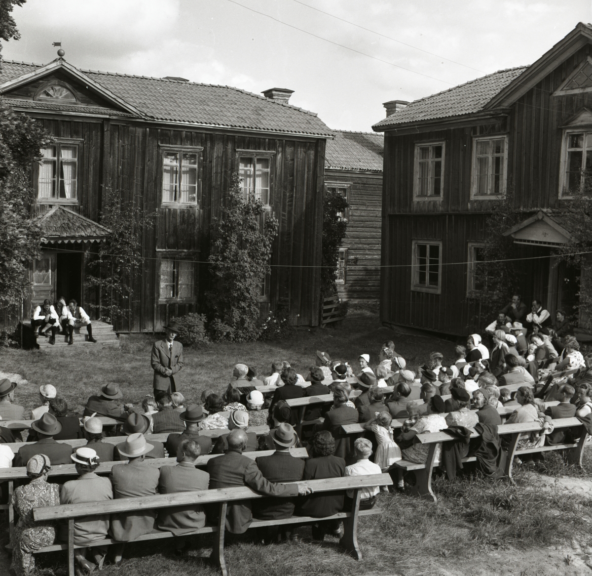 Framför två bostadshus vid hembygdsgården i Rengsjö håller en man tal inför publik, 28 augusti 1955.