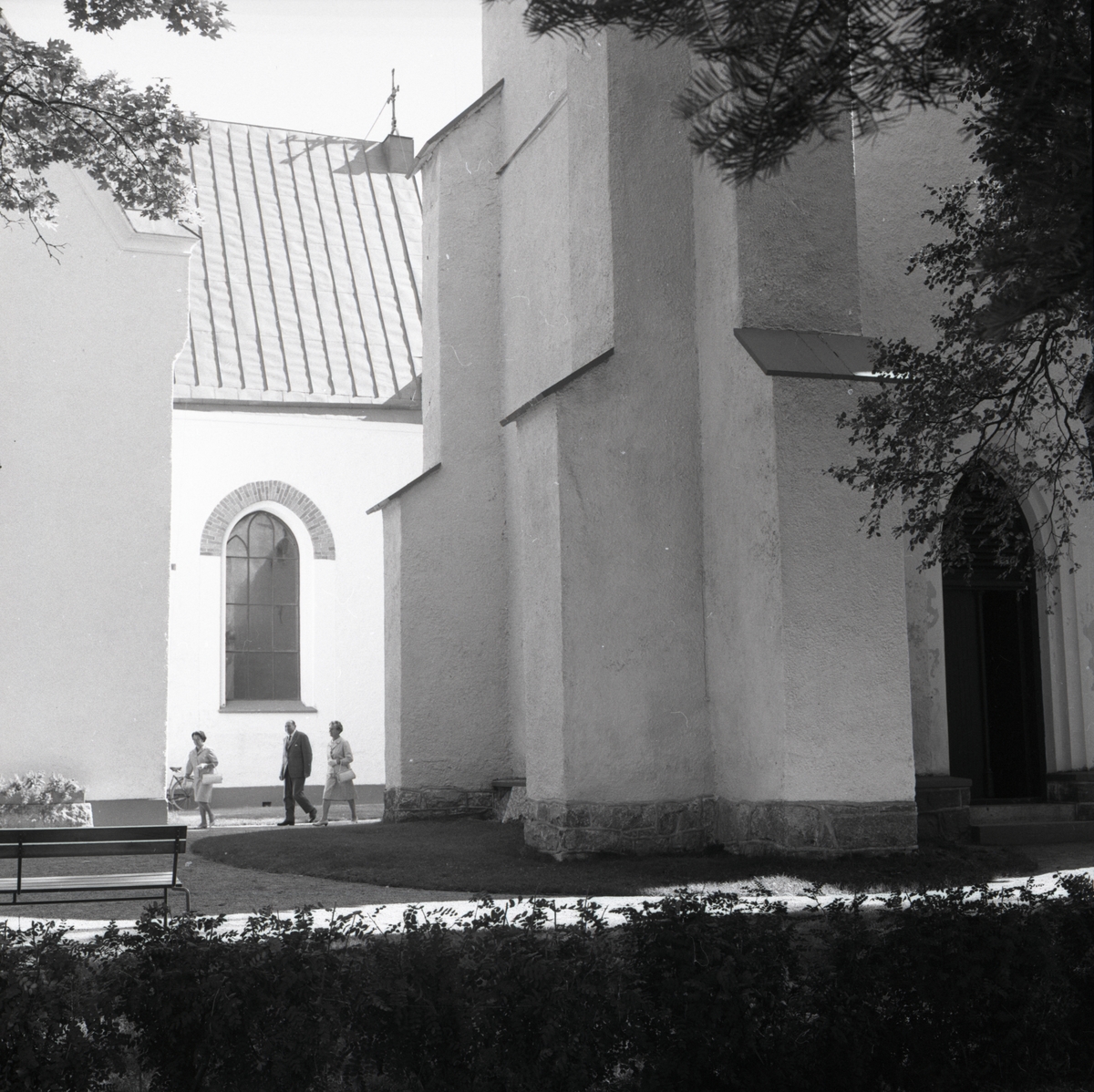 Tre personer vandrar över gårdsplanen intill Harmångers kyrka den 21 augusti 1966.