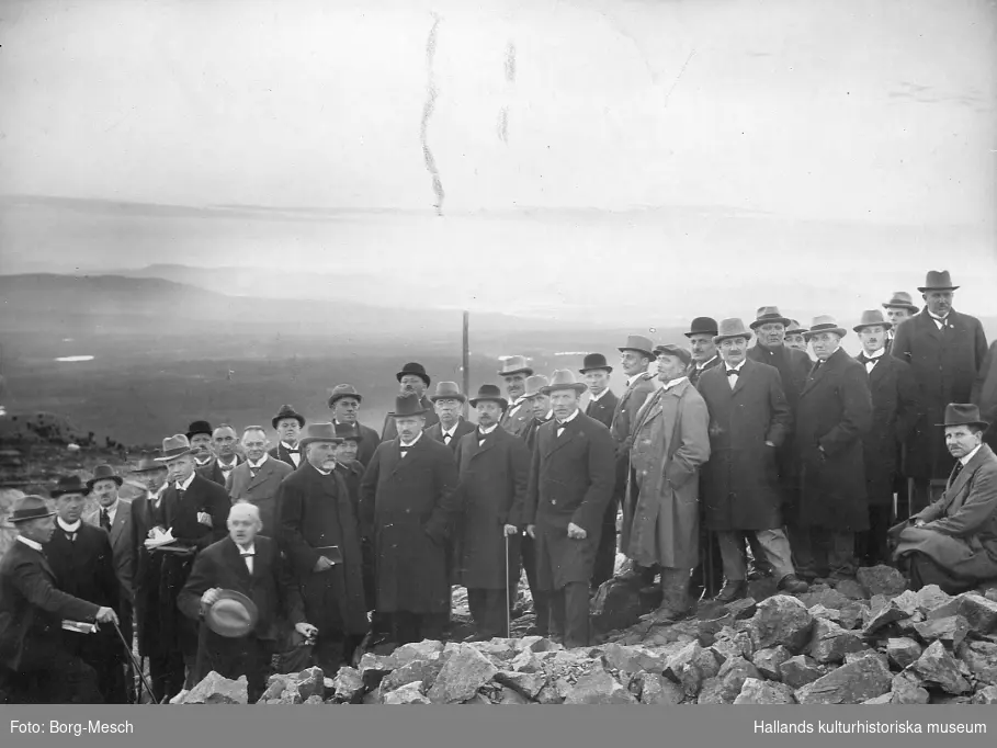 En stor grupp välklädda herrar i kostym, hatt och överrock, somliga med käpp, fotograferade bland stenar utomhus på en hög höjd.