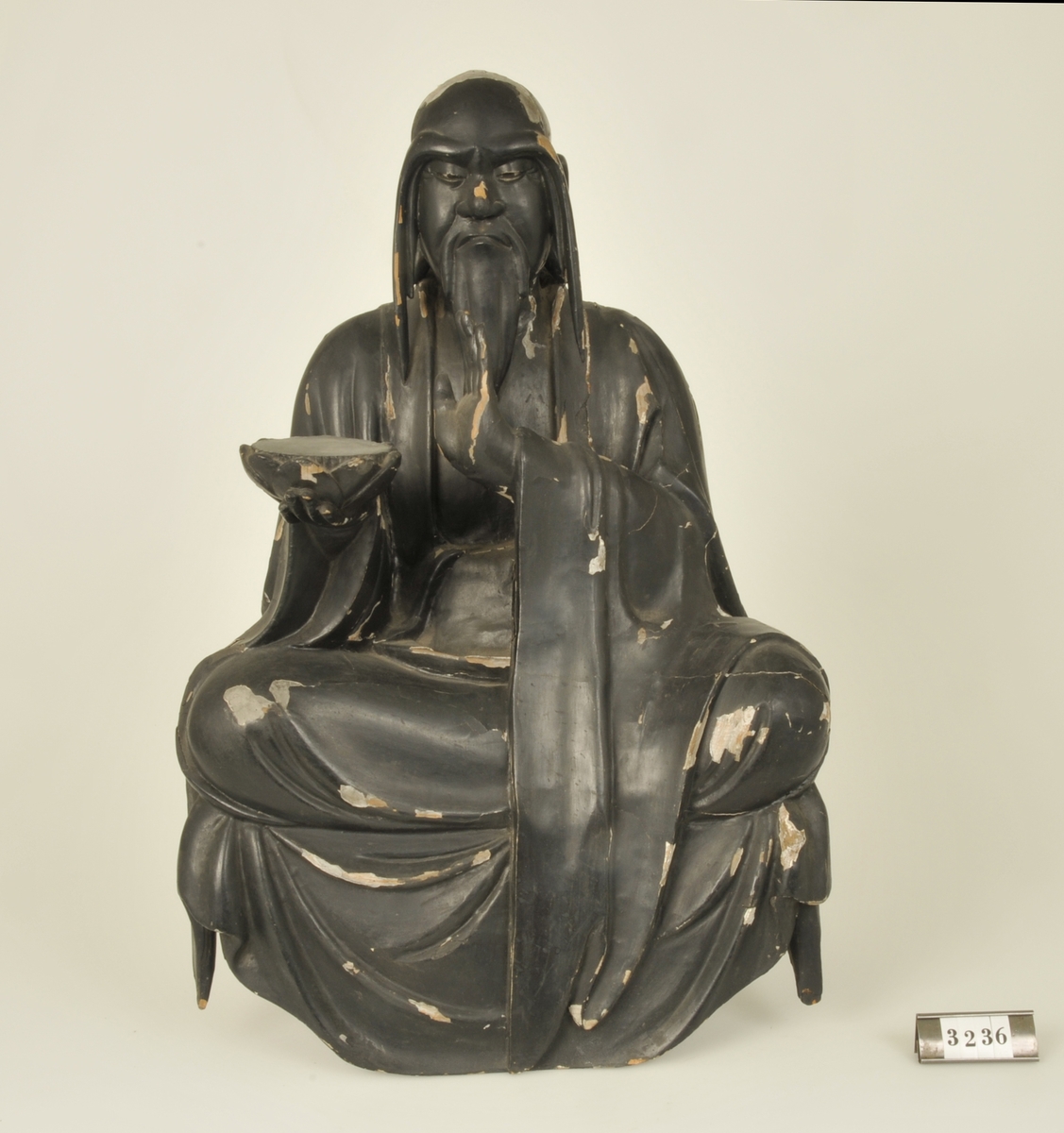 Träskulptur föreställande en man i sittande ställning.
Håller i höger hand en tom bricka. Målad i svart.

Har tillhört de Adelsköldska samlingarna.

