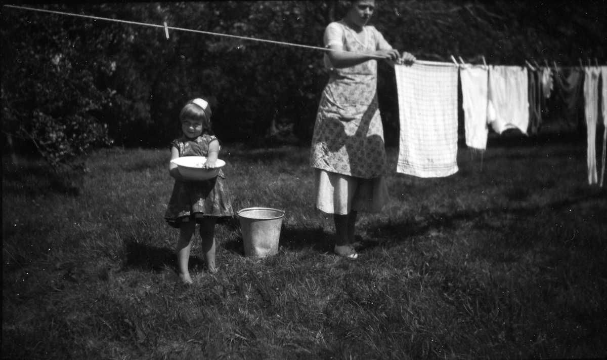 En kvinna och en flicka hänger tvätt på ett klädstreck, flickan håller i en balja.