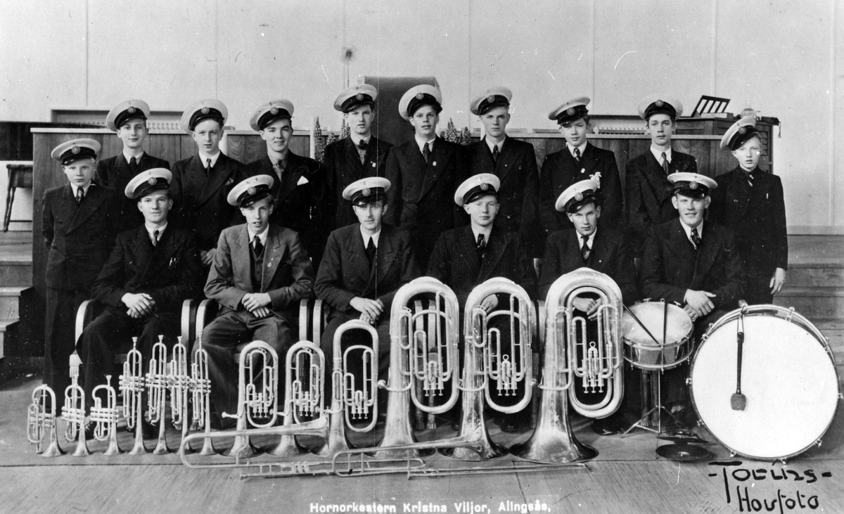 Gruppbild med 16 pojkar och män klädda i uniform från Missionkyrkans hornorkester - Kristna Viljor. Framför sig har de ställs sina musikinstrument i storleksordning.