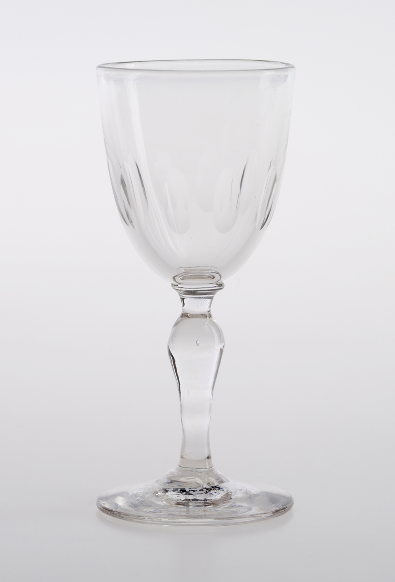 Et drammeglass laget av klart glass. Det er en langstrakt klokkeformet cupa med olivenslipninger i nedre del av klokken. Stetten er balusterformet.