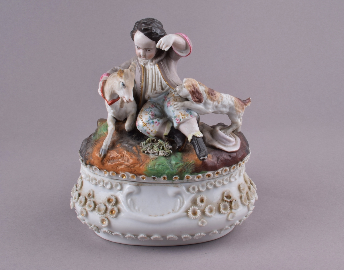 Oval skål med lokk. På lokket er en dekormalt figur av en gutt med en geit og en hund. Skålen er glasert og dekket av små uglaserte rosetter i relieff, noen med gulldekor i midten.