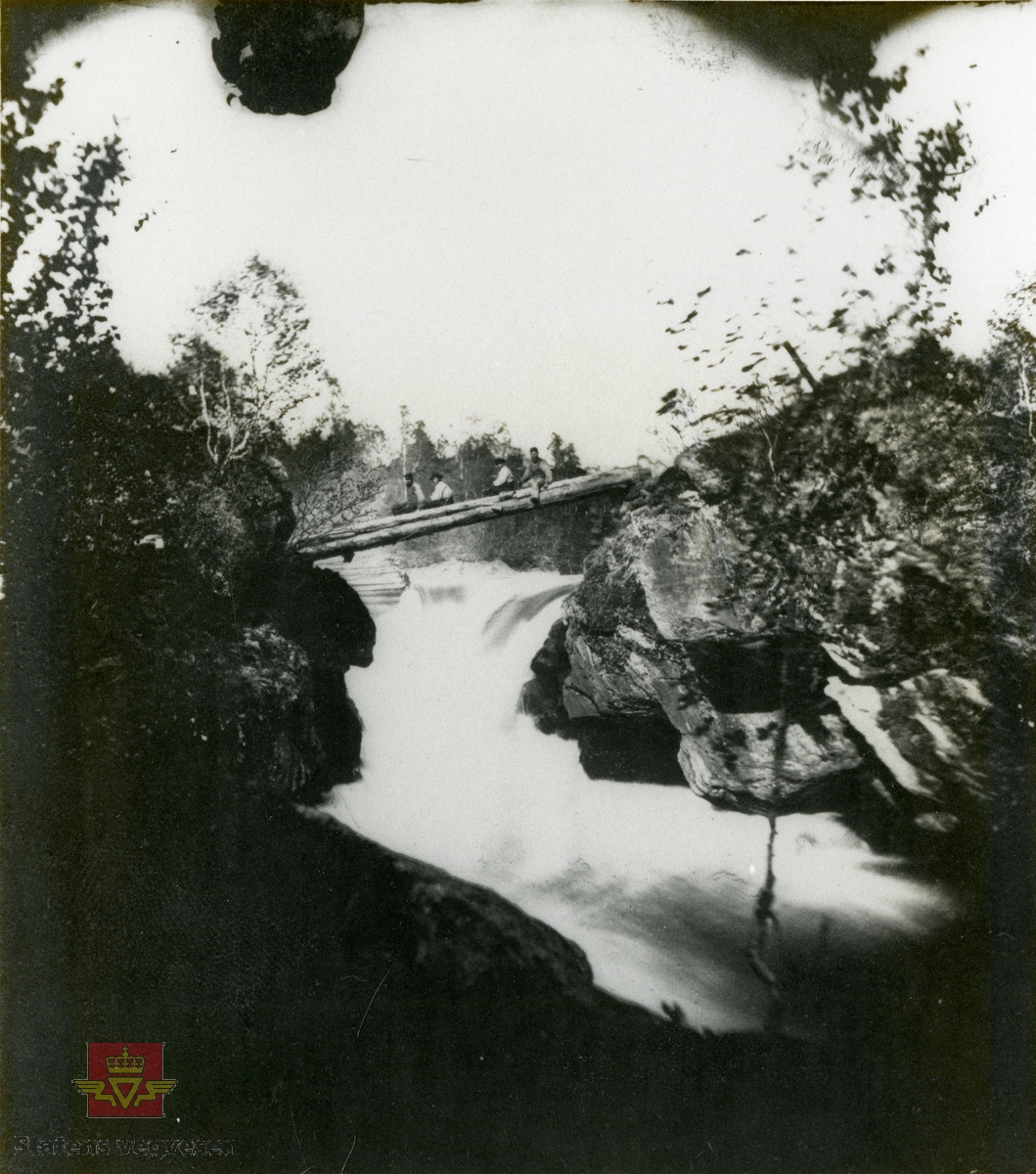 Personer sitter på stokkebru over elv i Hattfjelldal. 
Bilde tilhører OT OLsen arkivet på Universitetsbiblioteket.
OTO nr 95