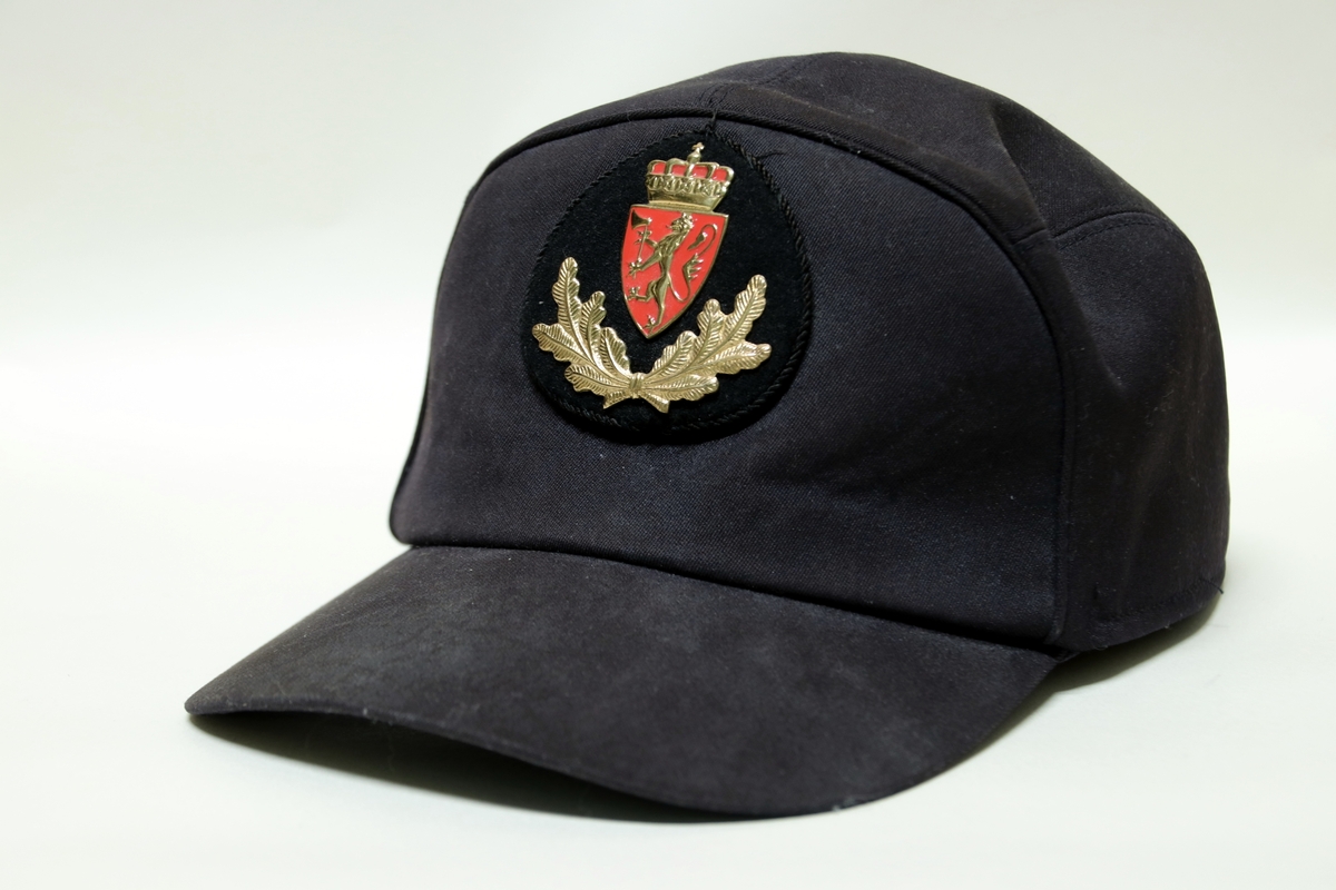Sort uniformslue med logo av norske riksvåpnet og eikeløv. Nedbrettbare ørevarmere.