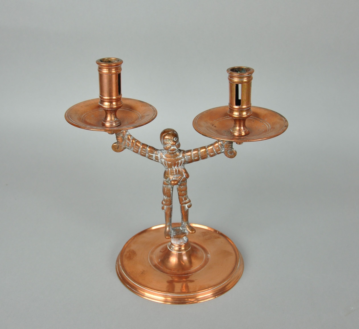 Lysestake av kobber. Staken er en mannsfigur som holder en dryppeskål i hver hånd. Mannsfiguren står på en rund sokkel.