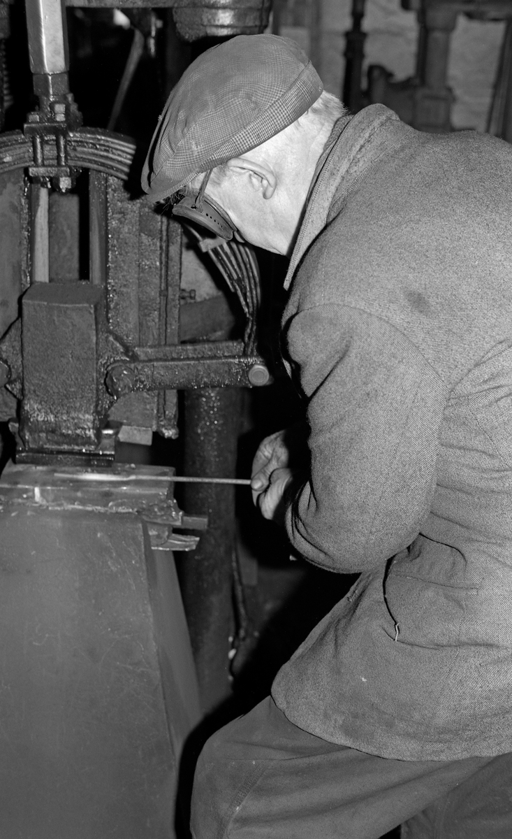Øksesmeden Oddmund Knudsen (1900-1983) i Mjøndalen i Nedre Eiker, fotografert ved maskinhammeren i Knudsen-smia, der han bearbeidet et glødende jernstykke.  Smeden hadde jakke på overkroppen og sixpencelue på hodet.  Framfor øynene hadde han briller med mørke glass.  Bildet ble tatt i 1974 da etnologen Åsmund Eknæs fra Norsk Skogbruksmuseum var i Knudsen-smia for å dokumentere arbeidet til den største leverandøren i merkeøkser til det norske skogbruket på 1900-tallet.