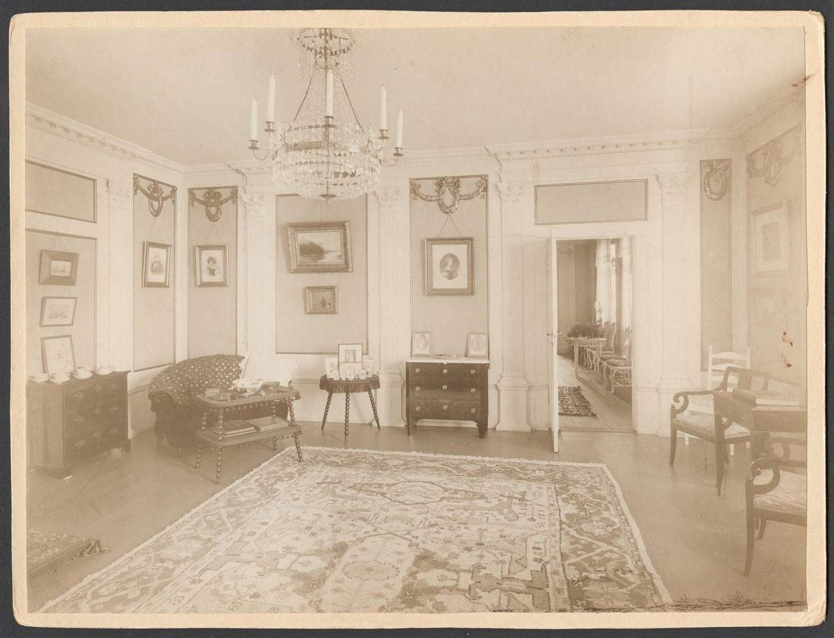 Bilden visar salongen (rum 114) i varvschefsbostället åren 1908-1912. Rummet är inred med klassicistisk interiör. Golvet är täckt med en stor matta och möblerna har arrangerats längs väggarna. Dörren står öppen och leder blicken vidare till den stora salen i första våningen (rum 112).