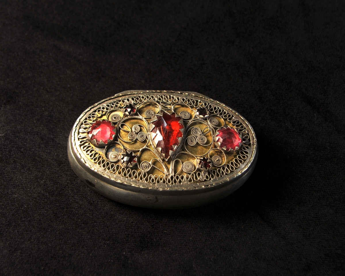 Oval, något buktande dosa av silver. Locket dekorerat med filigranarbete och 7 infattade stenar av rött glas. Invändigt förgylld. Stämplad invändigt i botten.