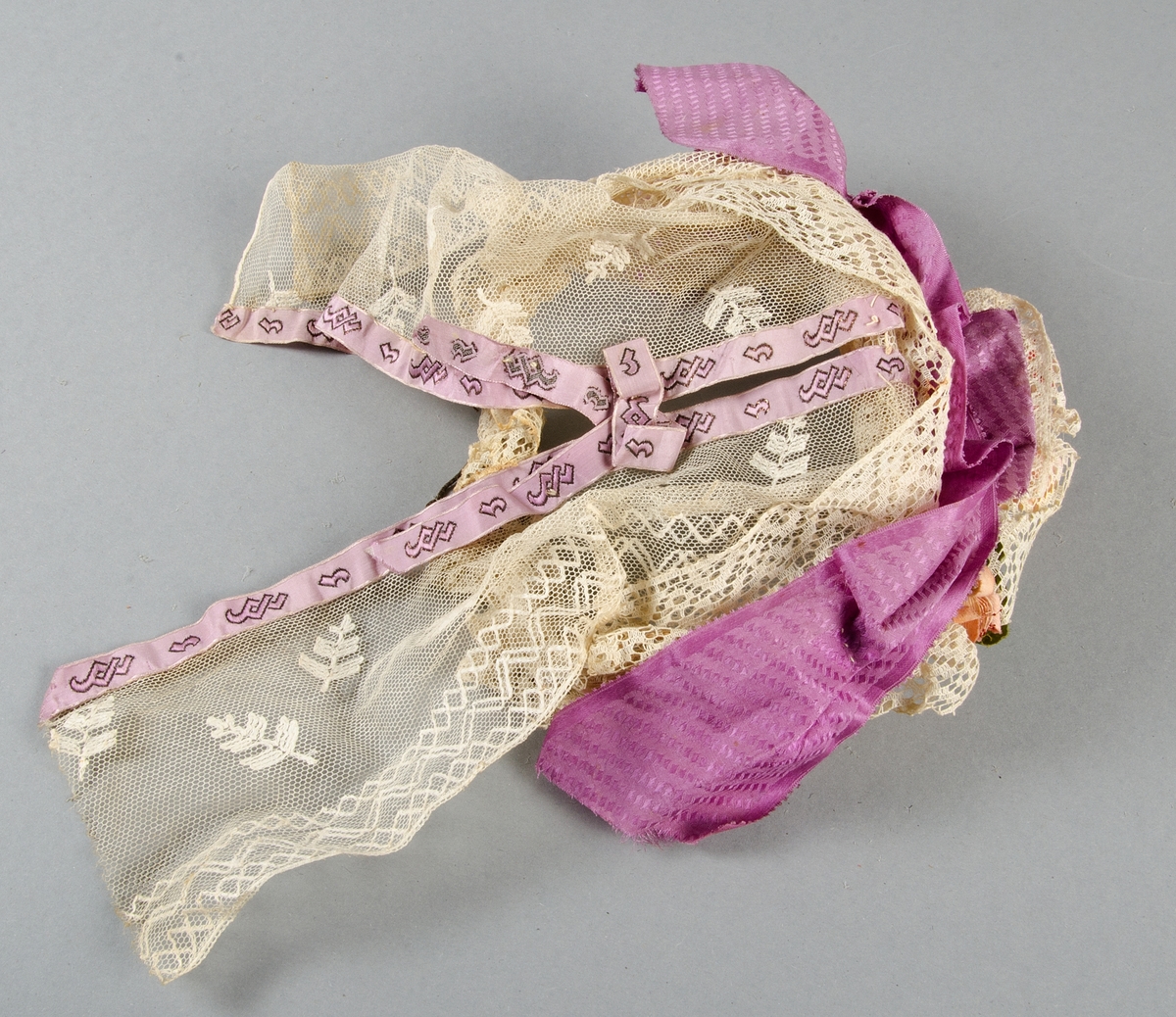 Hårklädsel av vita spetsar, mönstervävda lila band och rosa och röda tygblommor på stomme av mässing-tråd (?). Spetsarna, banden och tygblommorna smutsiga.