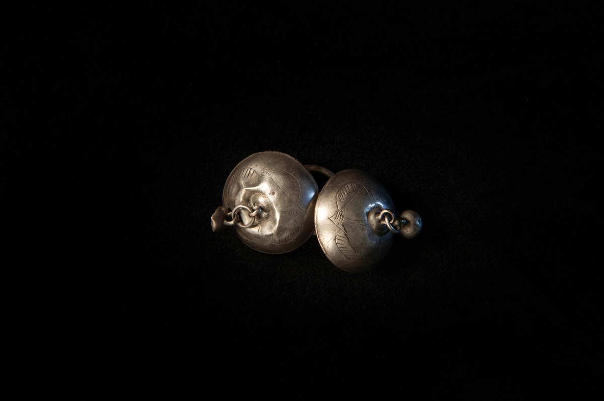Länkknapp av silver bestående av två kupiga knappar med kula, sammanbundna med ring. Enkel graverad streckdekor.