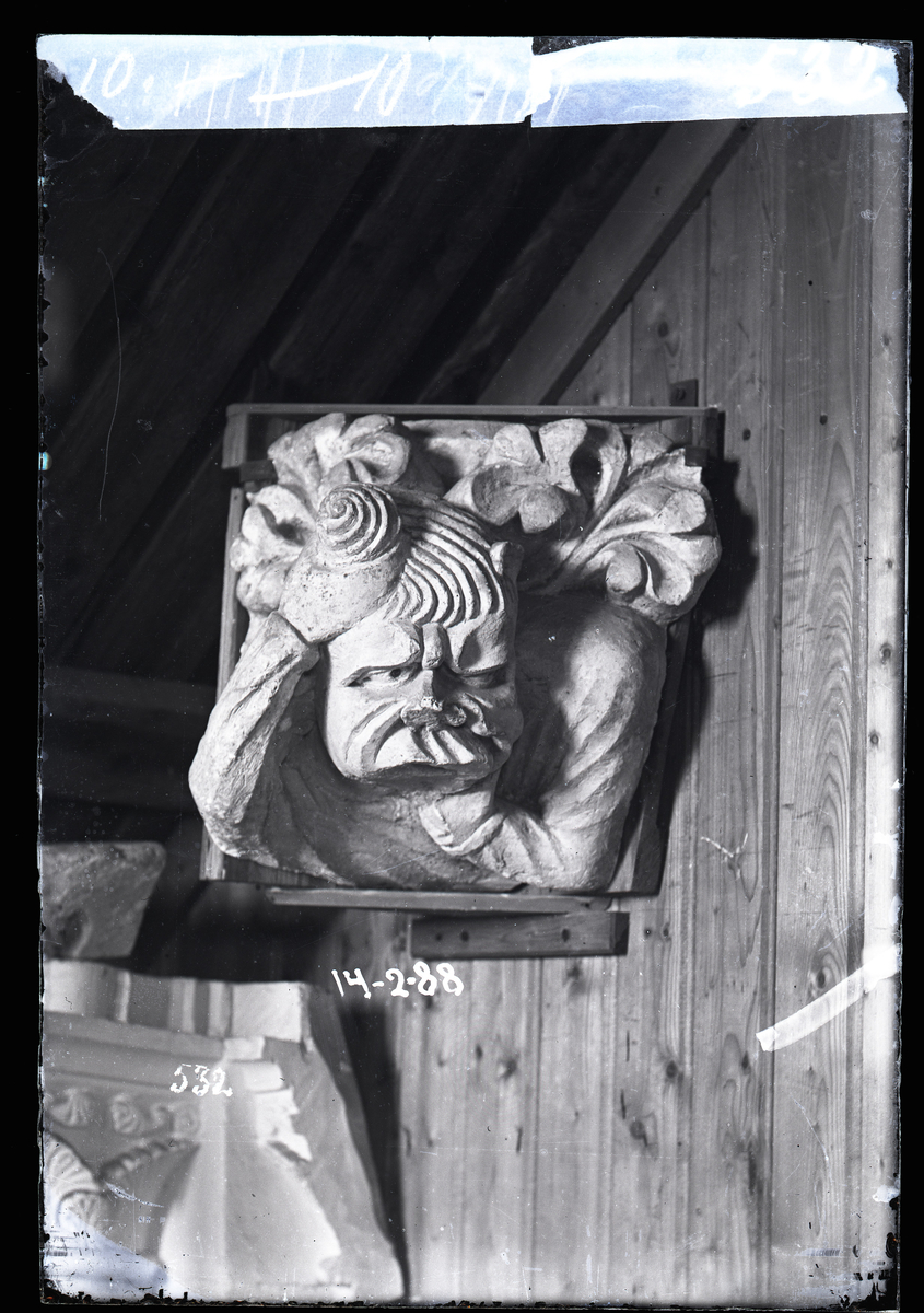 Gipsavstøpning av skulptur fra Nidarosdomen, plassert i et hjørne mellom tårnbuene innvendig. Person som holder seg til haken og i håret. Bladverk bak ryggen til personen.