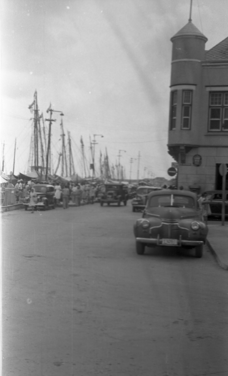 Gateparti. Kaien med seilbåter til venstre og bebyggelse til høyre. Biler og personer i gaten. Suderøy på vei til fangstfeltet.