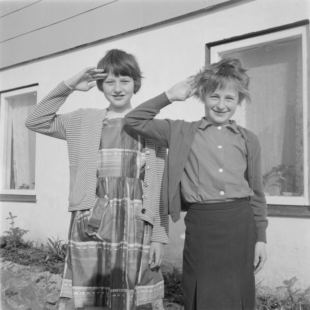 To unge jenter som gjør speiderhilsen?
Jenta til høyre er Lisbeth Olsen (nå Busch), født 19.07.1953. Det er trolig broren hennes, Erling Sven, som har tatt bildet. Han jobbet som journalist på avdelingskontoret til Helgeland Arbeiderblad i Brønnøysund. Bildet er tatt utenfor familiens hus i Nedre Flatåsvei i brønnøysund. Lisbeth og Erling er barn av Egil Schønning Olsen og Solveig Marie Olsen (født Busch). 
Jenta til venstre er trolig Lisbeths venninne, "Bjørgen".