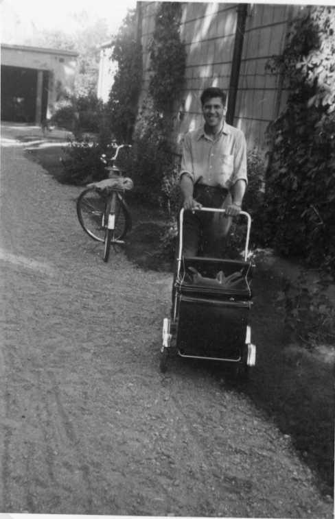 Man i skjorta med uppkavlade ärmar kör en barnvagn på en grusad uppfart. En cykel står parkerad bakom honom.