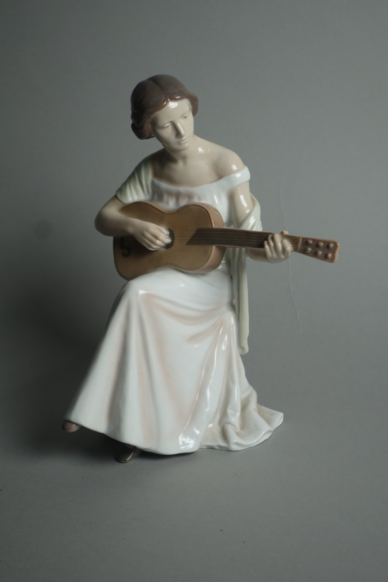 Porselensfigur formet som en gitarspillende dame iført lang hvir kjole og lysegult sjal. Hun sitter på en åttekantet tyrkisk taburett.