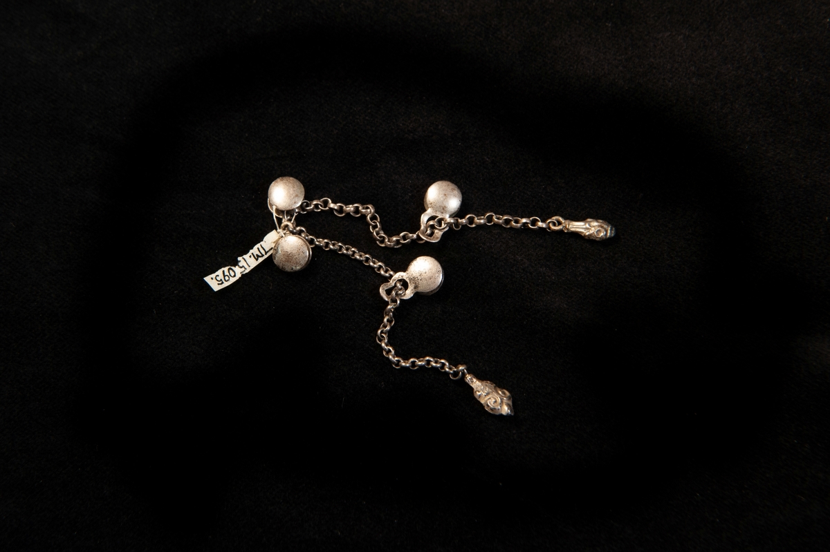 Knappar, en enkel kedja av silver med fyra små, runda så kallade stolplknappar. Kedjan avslutad med ovala små gjutna klumpar med enkel pressad dekor. Något otydliga stämplar (på knapparna).