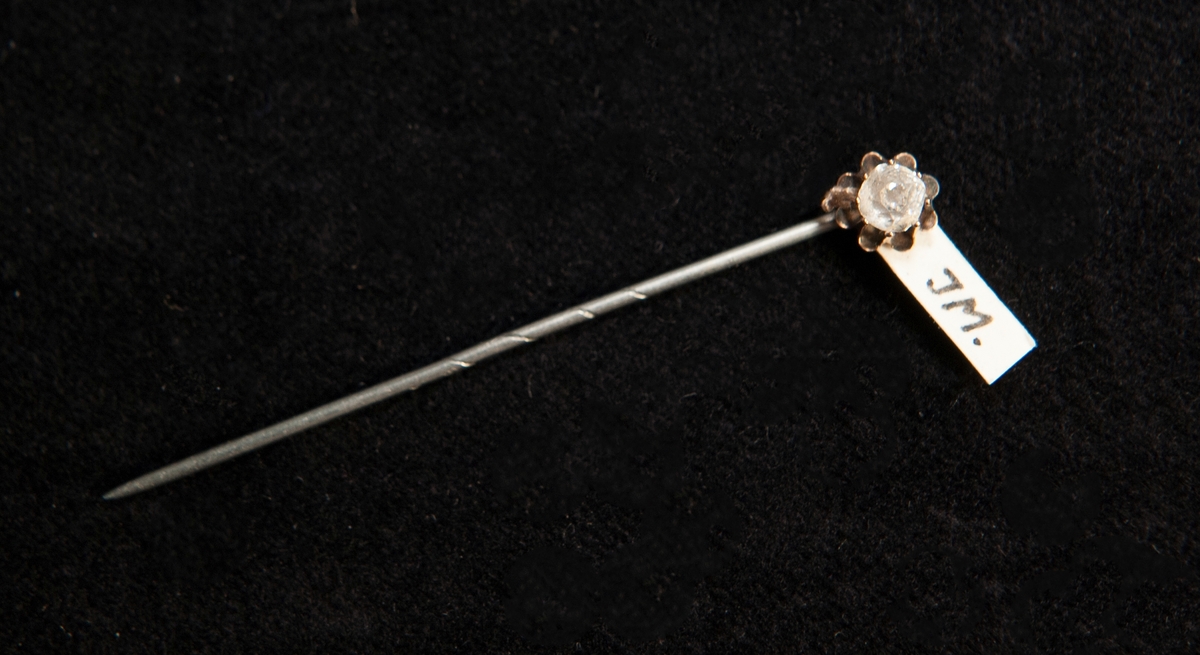 Kråsnål av silver med infattad klar glassten. En svårtydd stämpel finns längst upp på nålen.