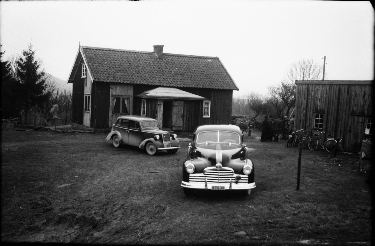 Två äldre bilar, "droskor", står parkerade på gården till en stuga; till vänster en Opel Regent ca 1937 och en Pontiac ca 1947 till höger. Vid uthusets vägg står några cyklar och i gattet mellan byggnaderna skymtar en folkklunga (se även bild EA1257). Till vänster om huset står en kärve till fåglarna.
