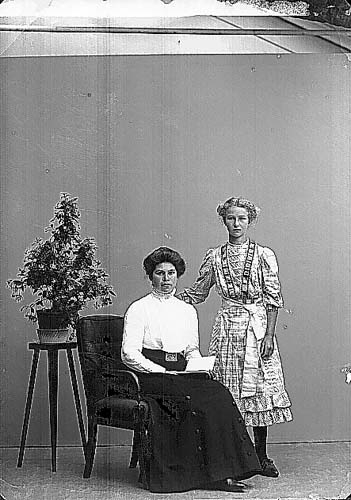 Pigan Ellen Blixt i ljus blus och mörk kjol sittande på en stol med en uppslagen bok i knät. Till vänster om en hög krukväxt på ett högt trebent blombord och till höger står Karin Johansson i klänning med veck och midjeband