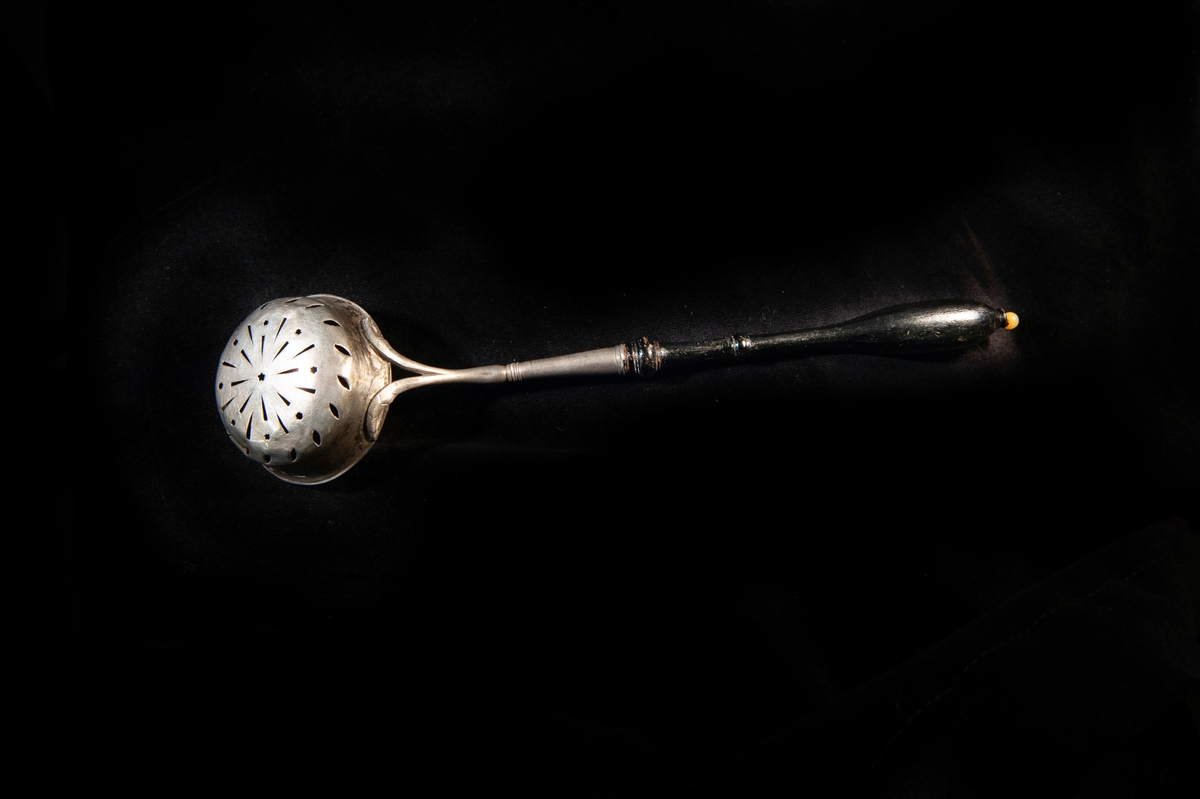 Strösked av silver, med djup perforerad skopa (med mönster av små stjärnor m.m.). Svarvat, svartlackerat träskaft med benknopp.
