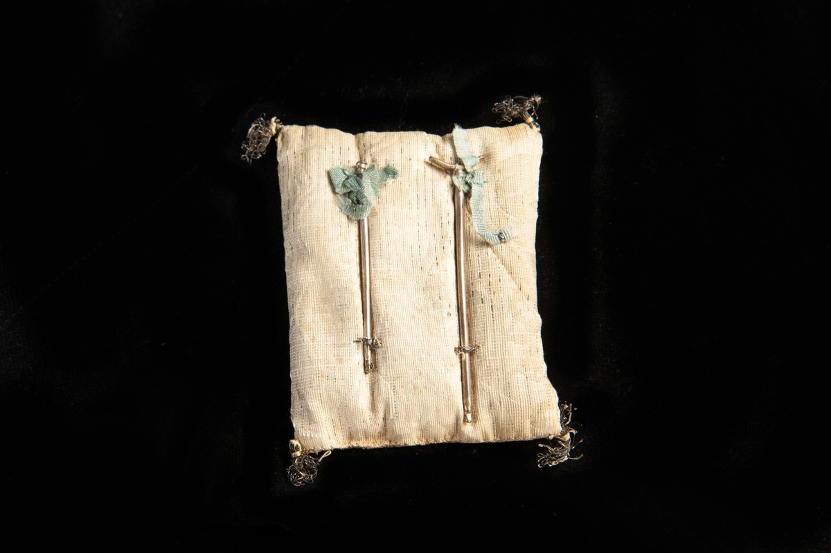 Käpp och krycka av silver monterat på potpouripåse (återanvänd textil med söm på både fram- och baksidan) med små tofsar i hörnen. Små blå rosetter är fästa vid käppen och kryckan.