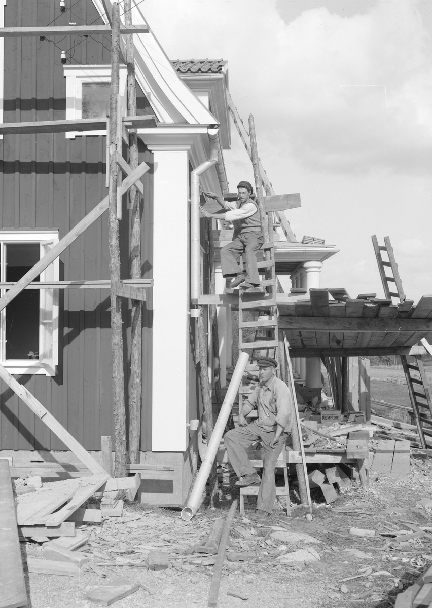 Ett nytt komministerbostället i Svinhult socken stod klart 1934. Här ses plåtslagarna Jonsson och Vallin montera husets sturrör.
De svaga ritsarna som kan anas i bildytan markerar hur fotograf Hultgren tänkt sig beskära bilden.